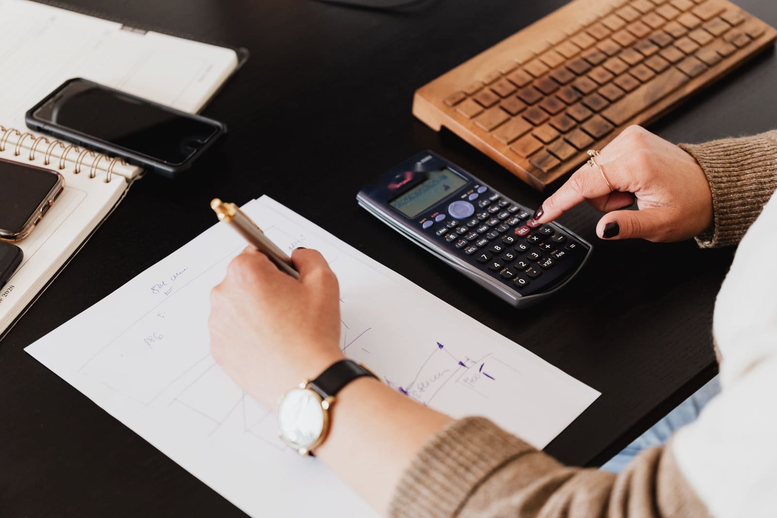 Hands working a calculator and sketching figures on a sheet of paper at a dark desk