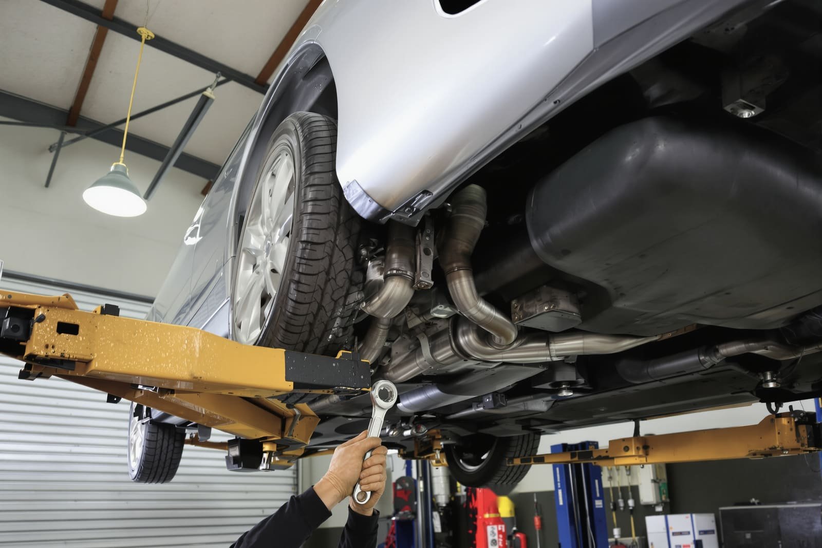 Looking up at the underside of a car on a two-post lift in a repair bay, a gloved hand holding a wrench to the exhaust
