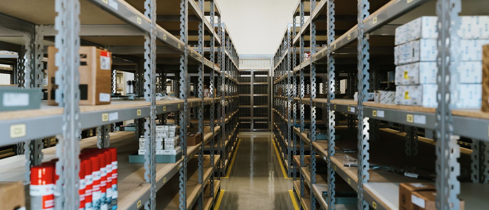 A long aisle between grey steel shelving stocked with cartons and labelled parts, a polished concrete floor with yellow lane lines