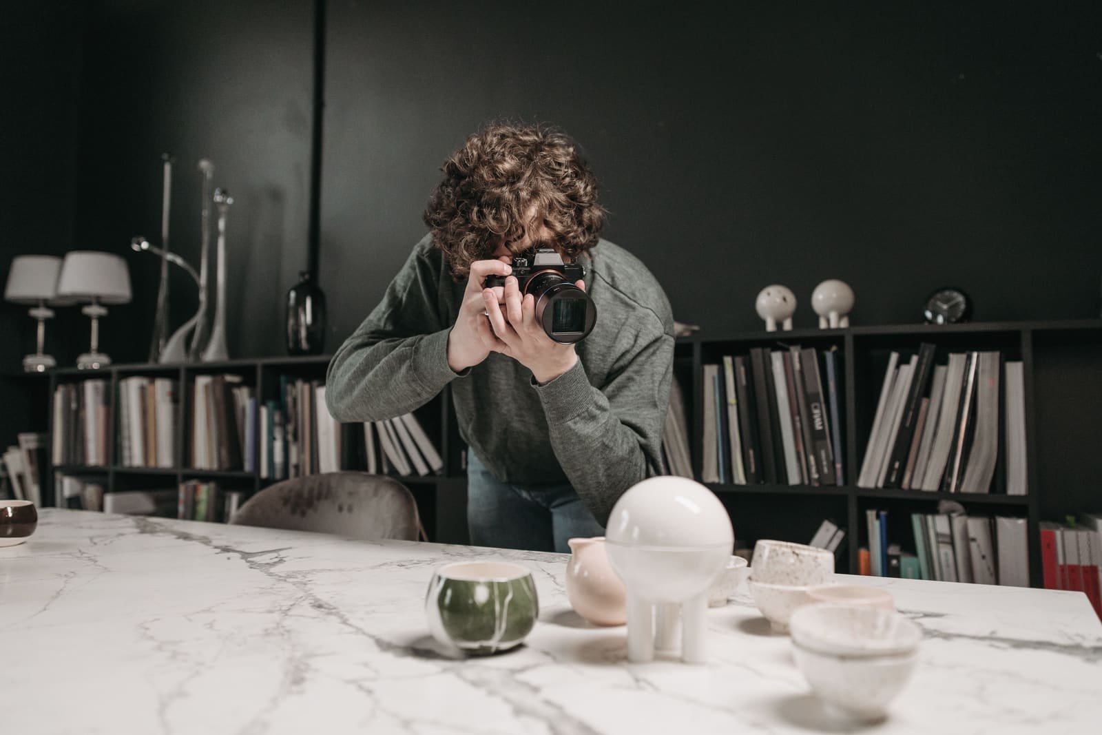 Someone leaning over a marble table to photograph handmade ceramics, bookshelves behind