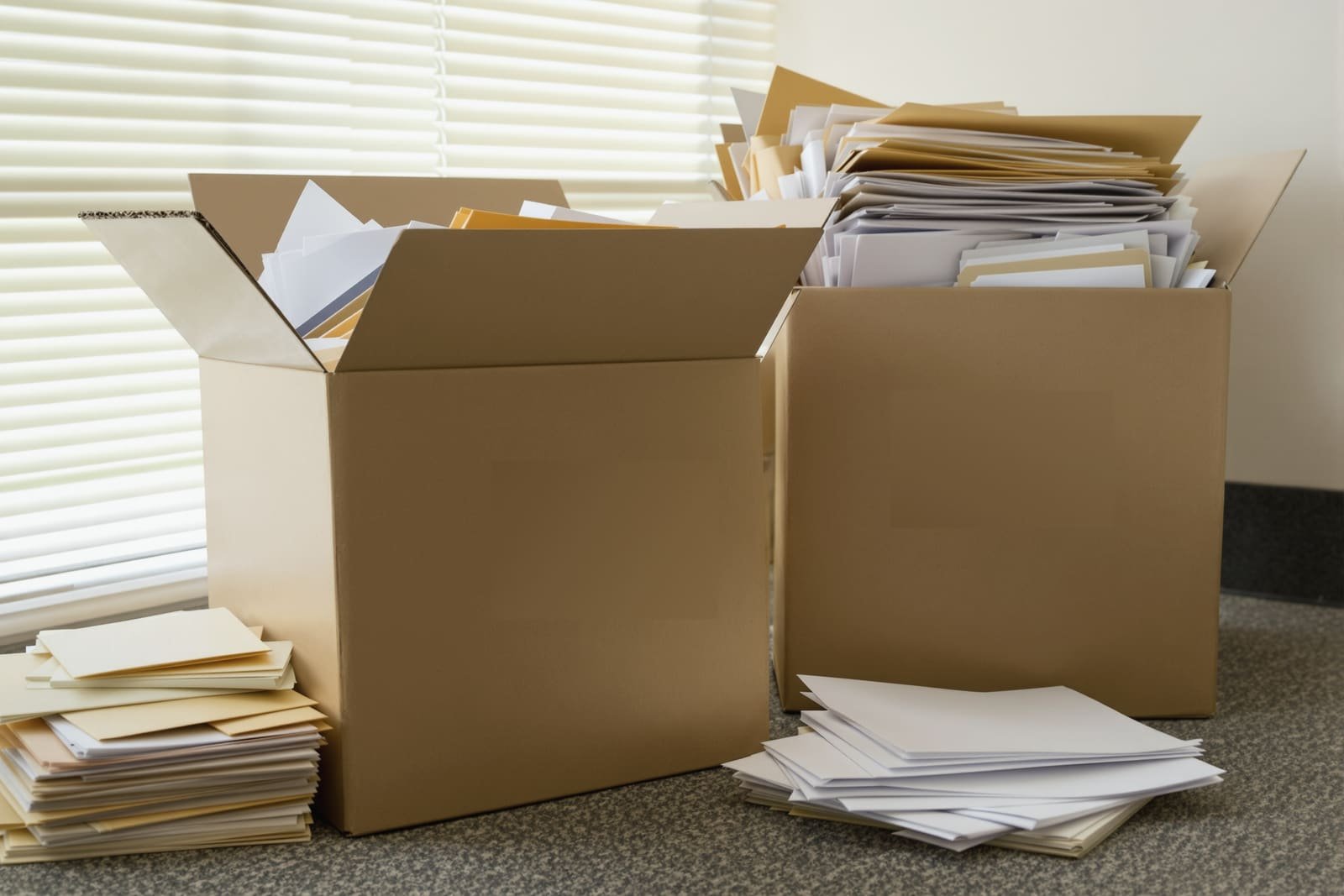 Two cardboard boxes overflowing with paper and folders on an office floor, more stacks beside them, blinds behind